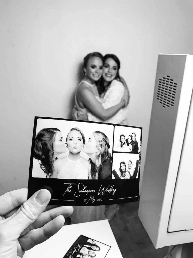 A hand holds a black and white wedding photo card in front of two guests embracing.